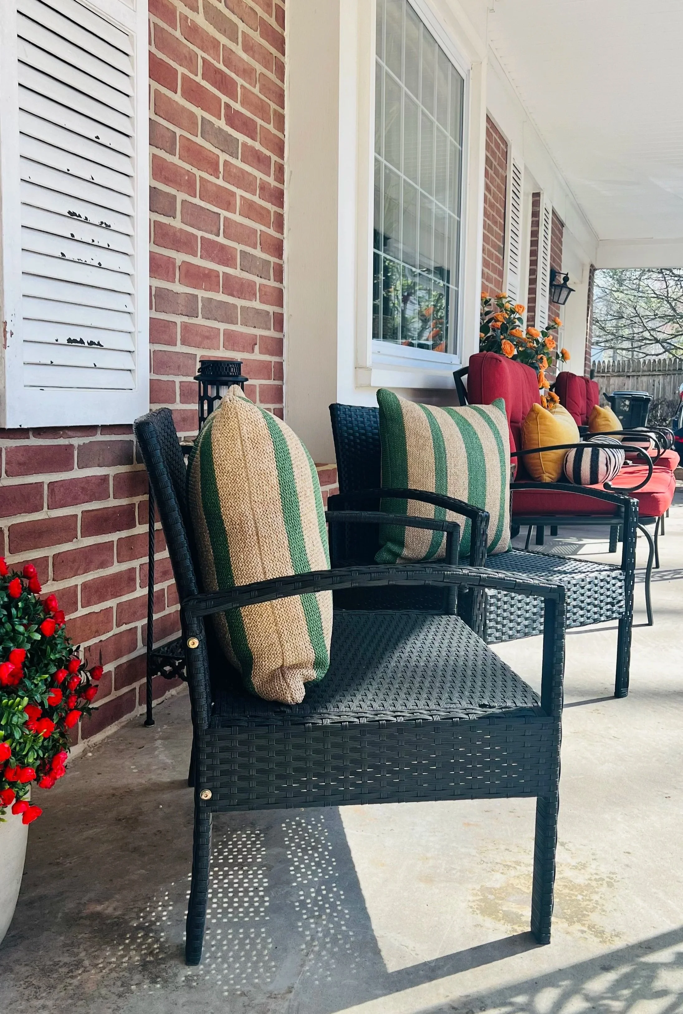 Covered front porch with red cushioned seating, decorative pillows, potted plants, and orange pumpkins, with a garden in the background.