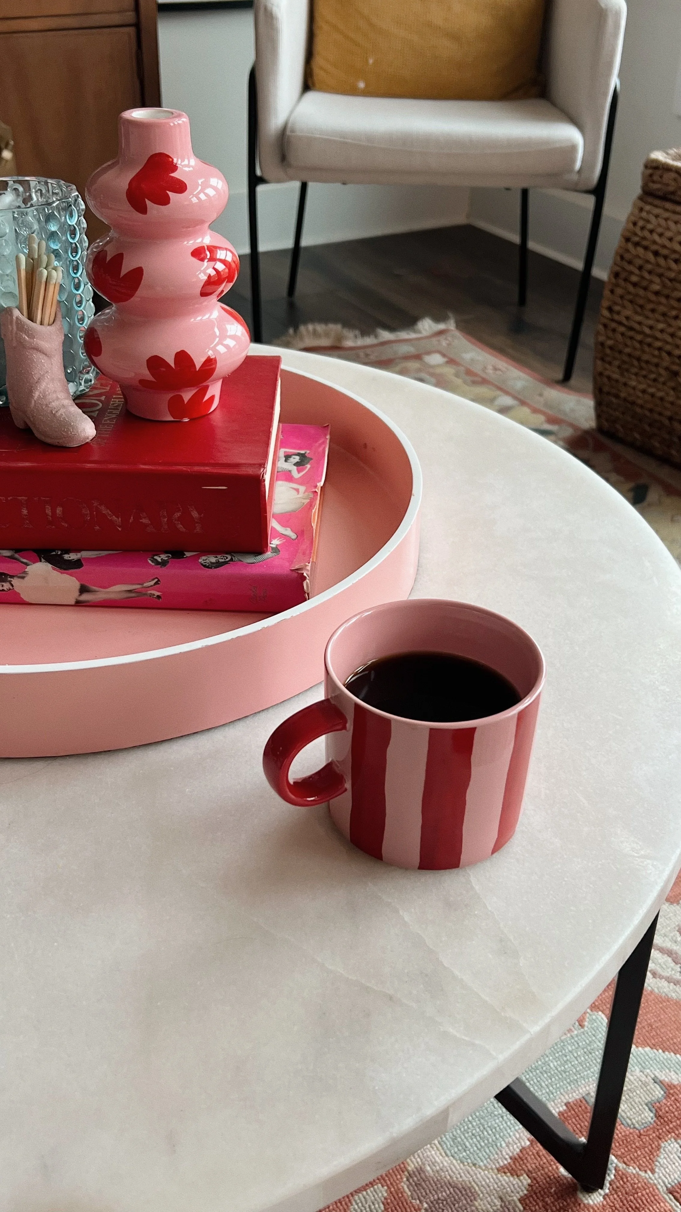 A pink and red striped mug filled with black coffee on a white table, with a pink decorative tray holding a pink and red vase, pink books, and a small pink boot-shaped container.