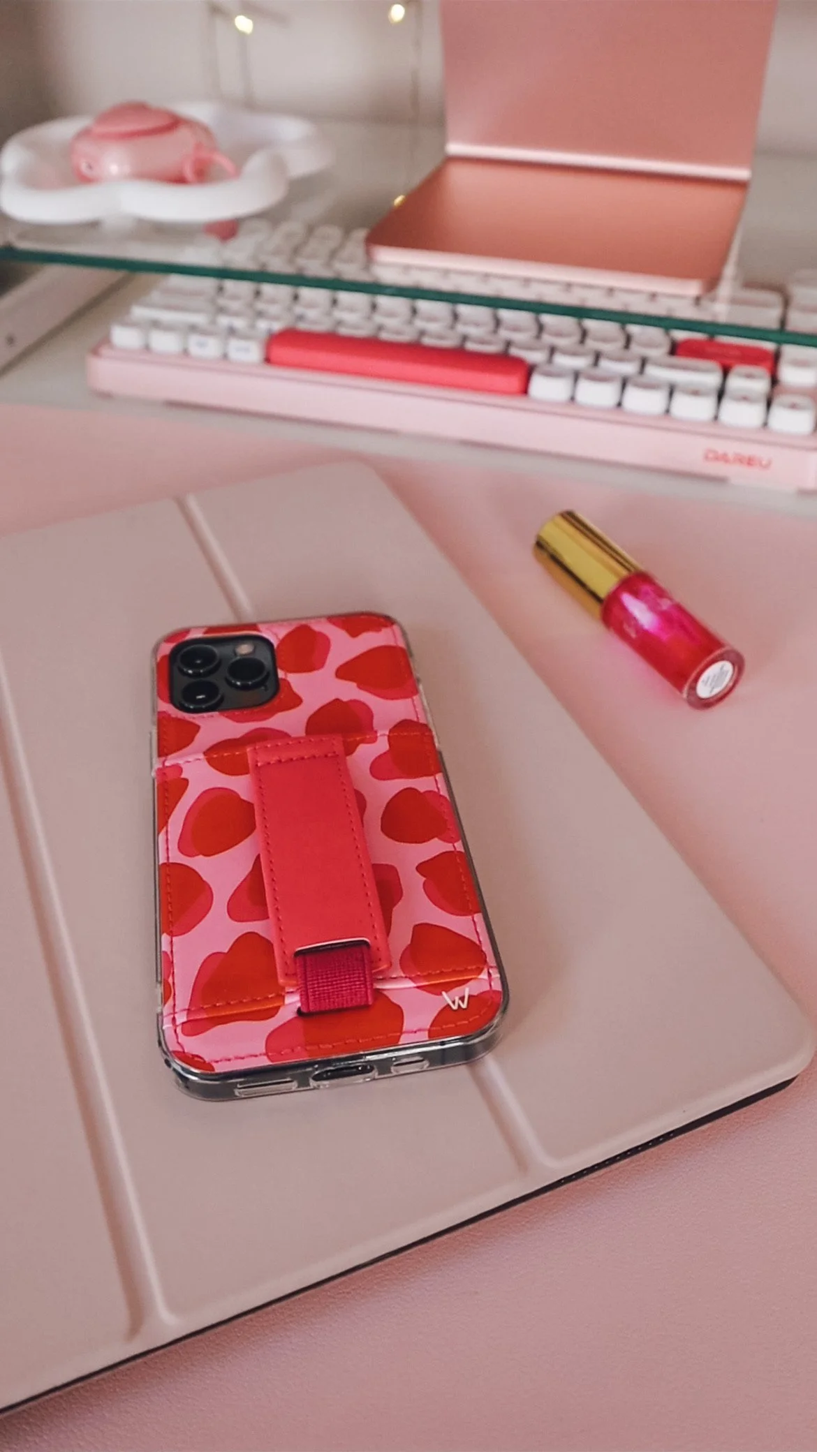 A pink smartphone with a red heart pattern and a pink strap on it, placed on a pink surface. In the background, there is a pink and white keyboard, a pink notebook, a small pink object on a white dish, and a pink and gold lipstick tube.