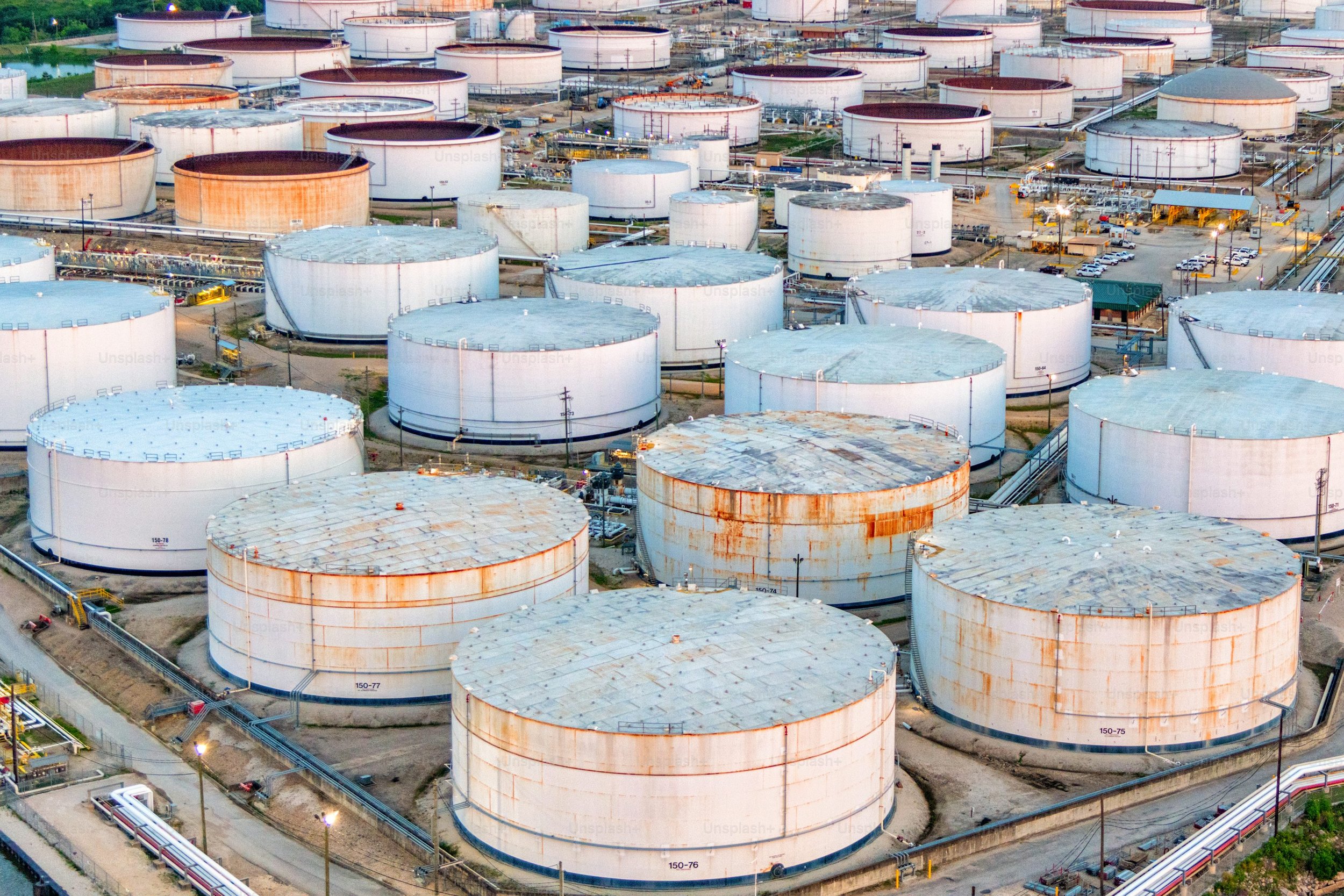 Aerial view of numerous large, cylindrical storage tanks at an industrial oil refinery or storage facility, some with rust and others painted white, with pipelines and vehicles around them.