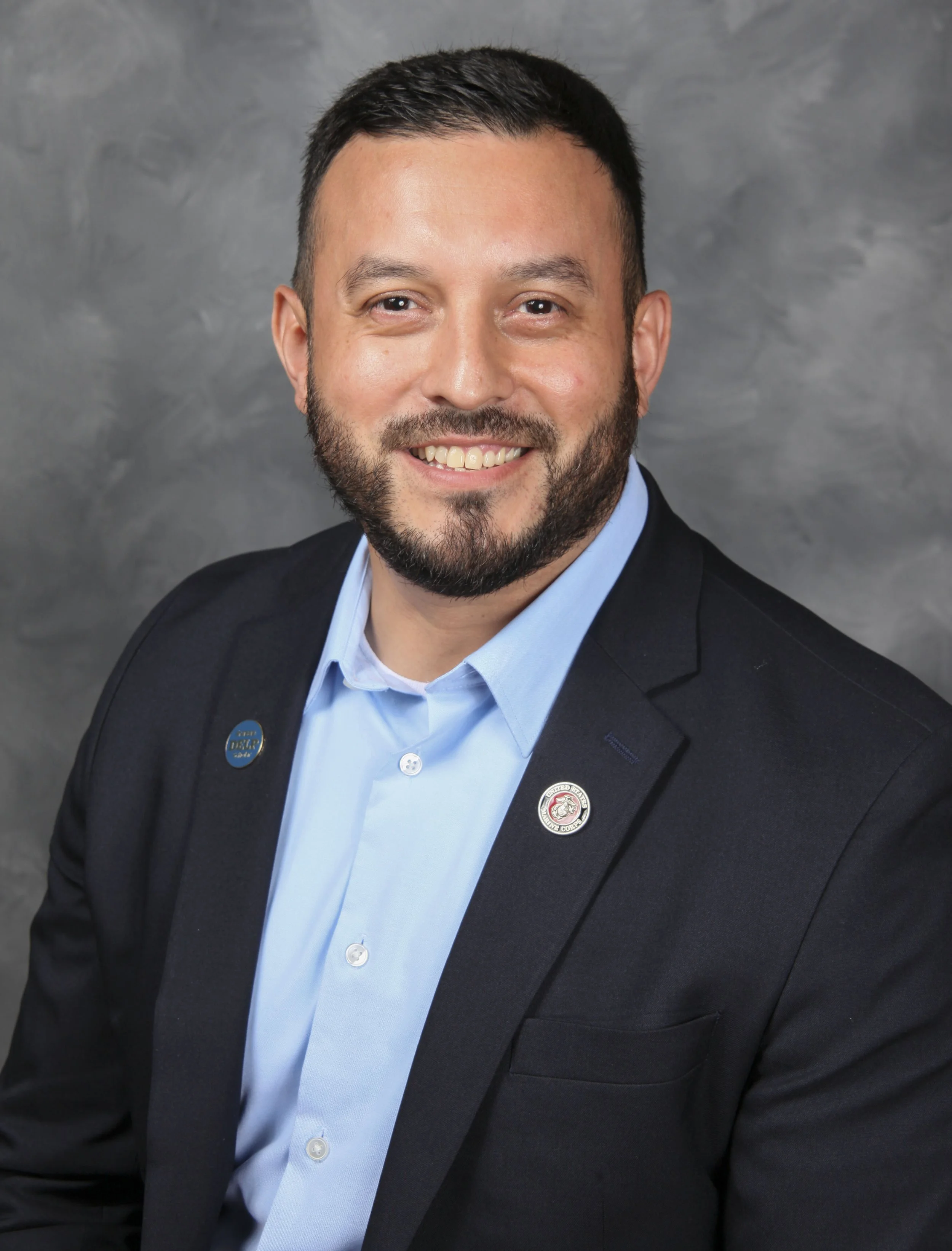 A man with short dark hair and a beard, smiling, wearing a black suit jacket and light blue dress shirt, with pins on his lapel, against a grey background.