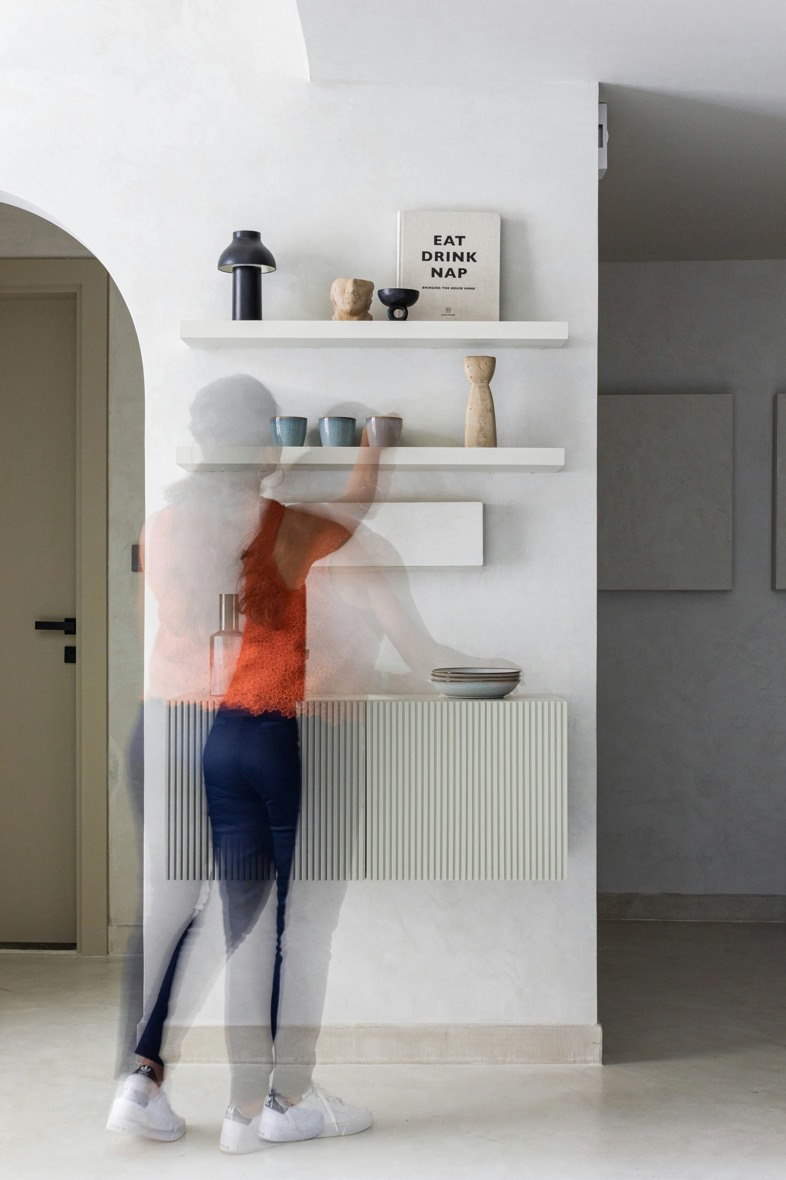 A woman in an orange top and blue jeans reaching for items on white wall-mounted shelves in a modern, minimalistic interior with a neutral color palette.