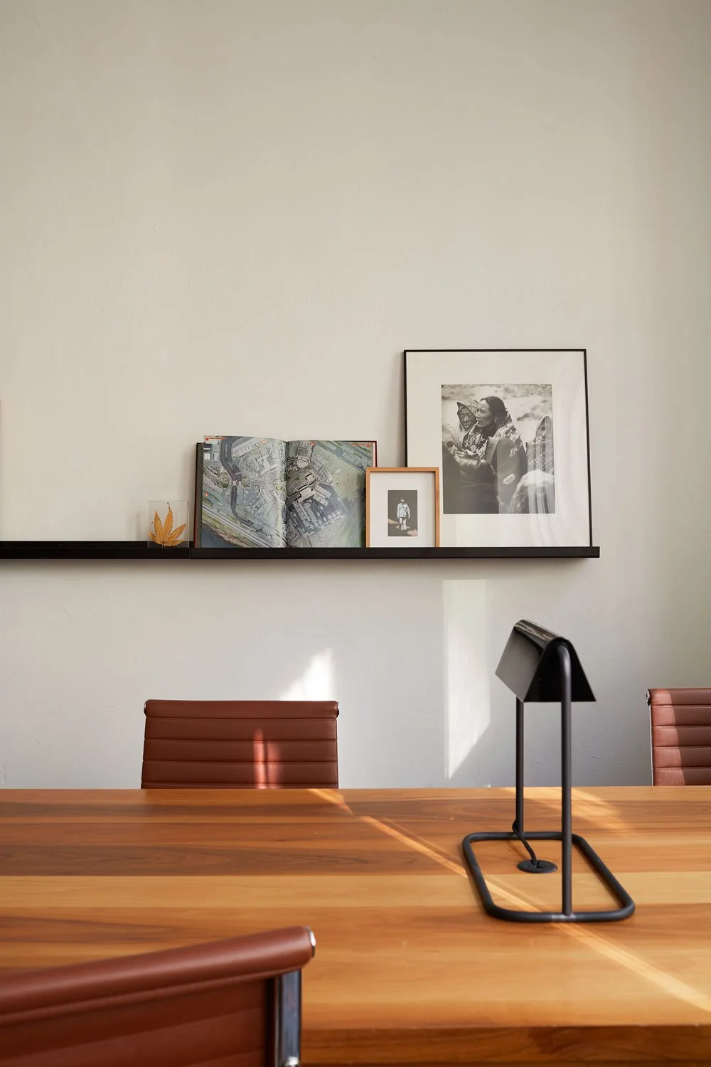 Modern office interior with brown leather chairs, wooden table, minimalist black desk lamp, and decorative shelf with framed pictures, open book, and leaf display.