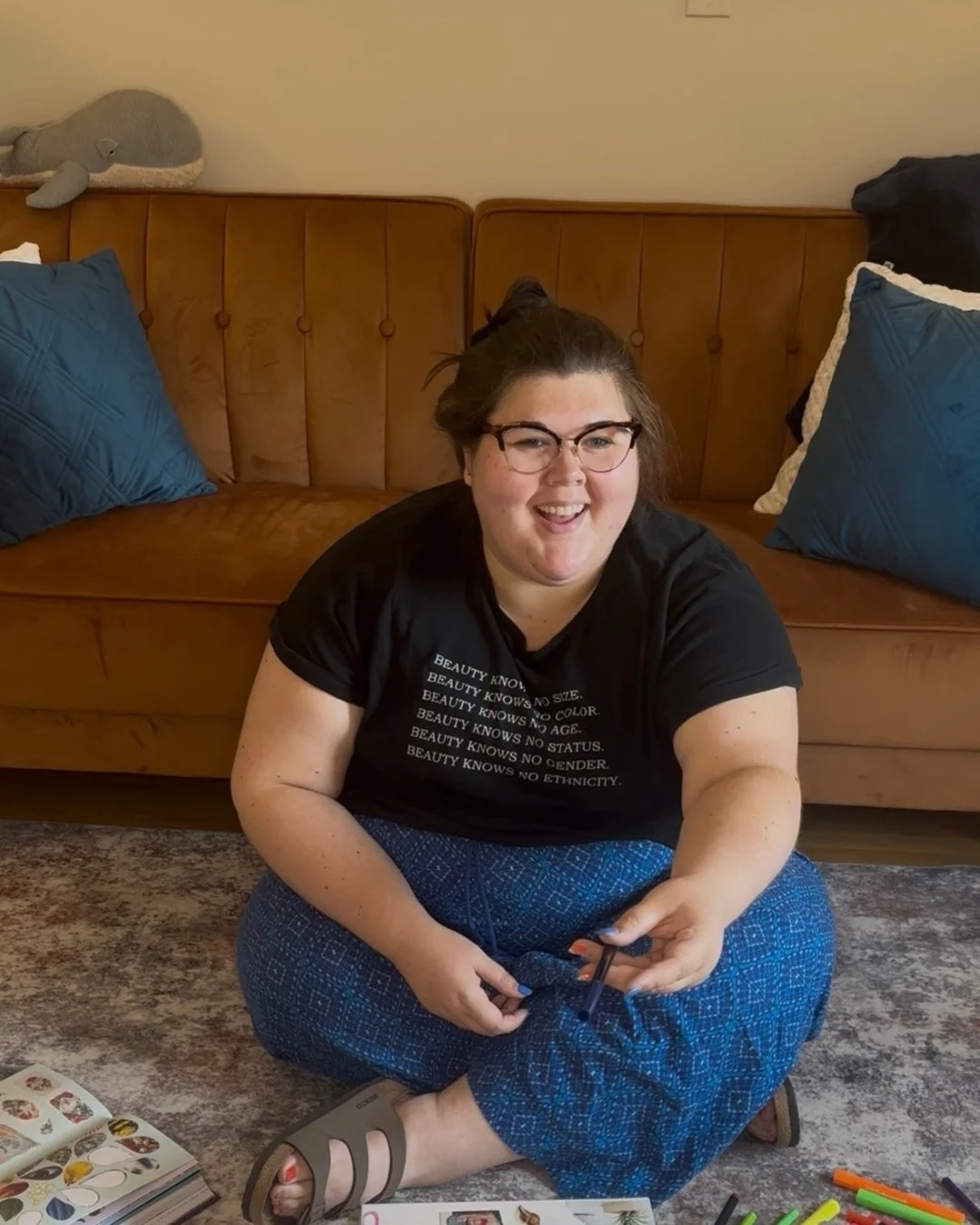 Smiling woman wearing glasses and a black T-shirt sits cross-legged on the floor in front of a brown leather sofa, holding scissors and surrounded by coloring supplies.