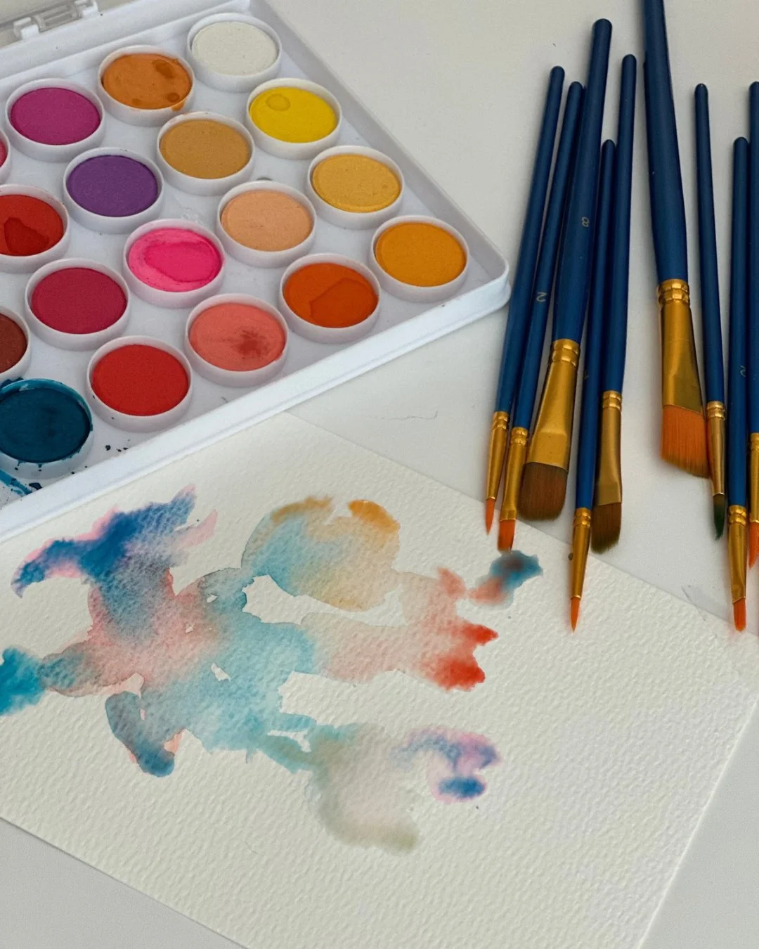 Watercolor paint palette, blue and gold paintbrushes, and colorful watercolor swatches on paper