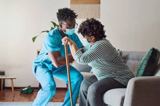 A physical therapist assisting a woman with a knee exercise at home, both wearing face masks.