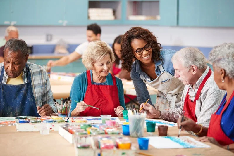 A diverse group of elderly people and a young instructor engaging in a painting class at a community center, surrounded by art supplies.