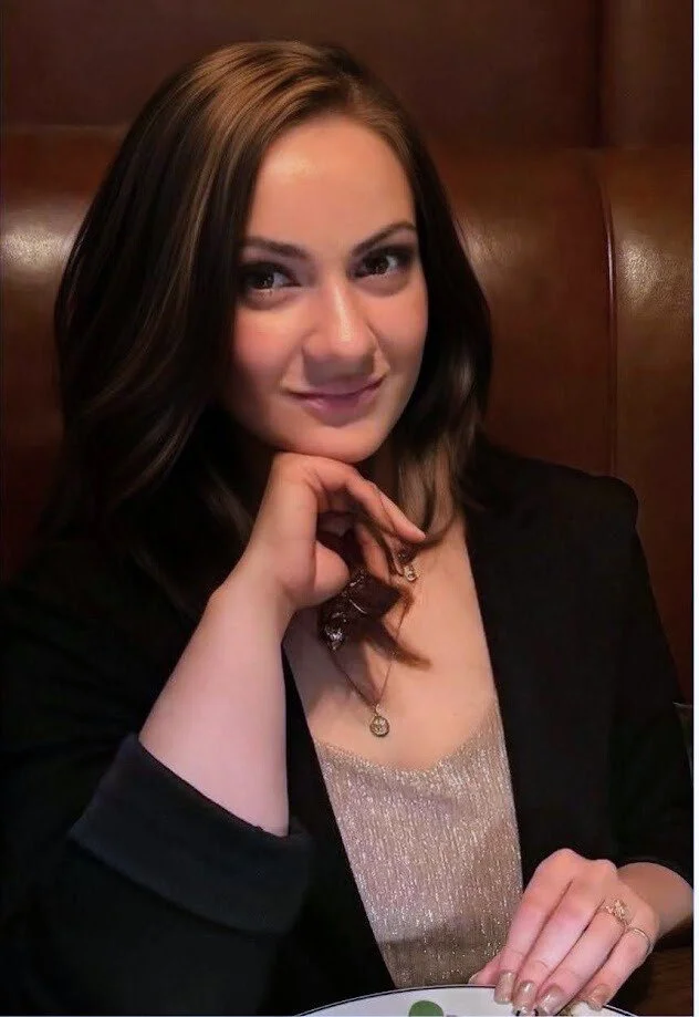 A young woman with brown hair styled in loose waves, wearing a black blazer and a shimmery top, sitting in a leather booth.