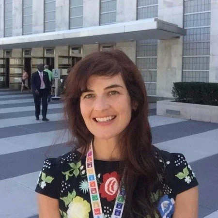Woman smiling outdoors in front of a modern building with glass windows, wearing a colorful lanyard and floral-patterned dress.