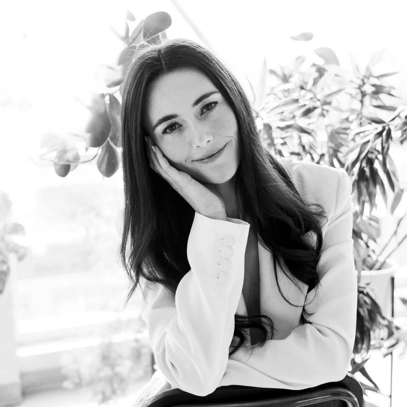 Black and white portrait of a smiling woman with long hair, resting her head on her hand, wearing a white blazer, with plants in the background.