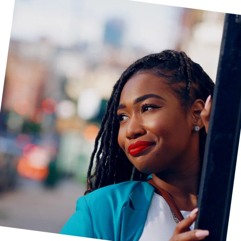 Smiling person with braided hair and red lipstick, wearing a turquoise jacket, looking into the distance.