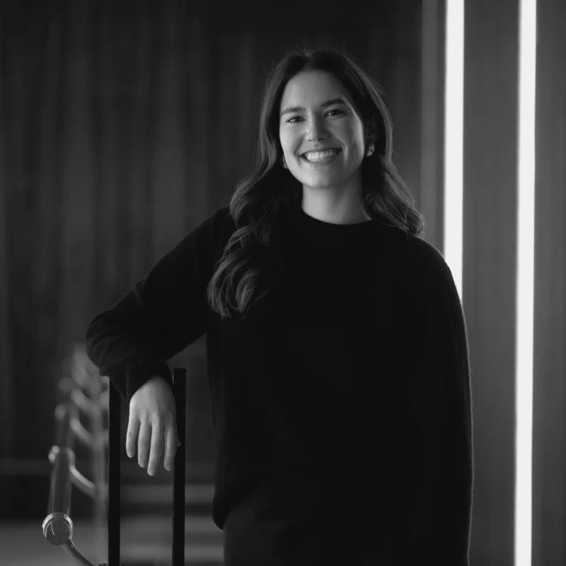 Black and white portrait of a person smiling, wearing a dark sweater, with long hair. They are standing indoors, resting their arm on a railing.