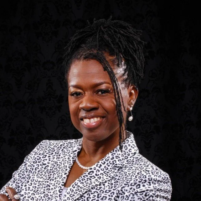 Smiling woman with dreadlocks wearing a patterned blazer against a dark background.