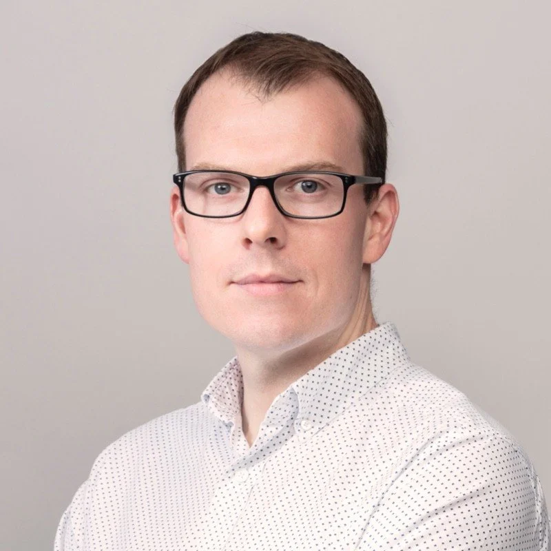 Man wearing glasses and a white button-up shirt with small black dots, against a plain background.