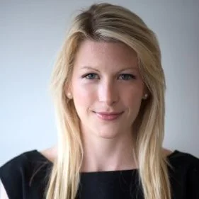 Portrait of a woman with long blonde hair, wearing a black top, smiling slightly against a plain background.