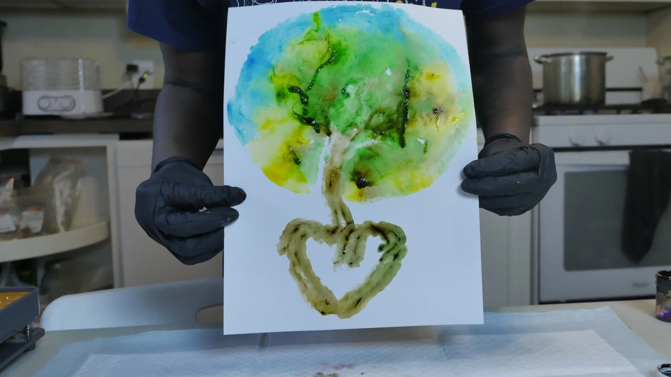 A person with long dreadlocks and earrings working at a kitchen counter with various artwork, plants, and display materials, and a window showing a view of trees outside.