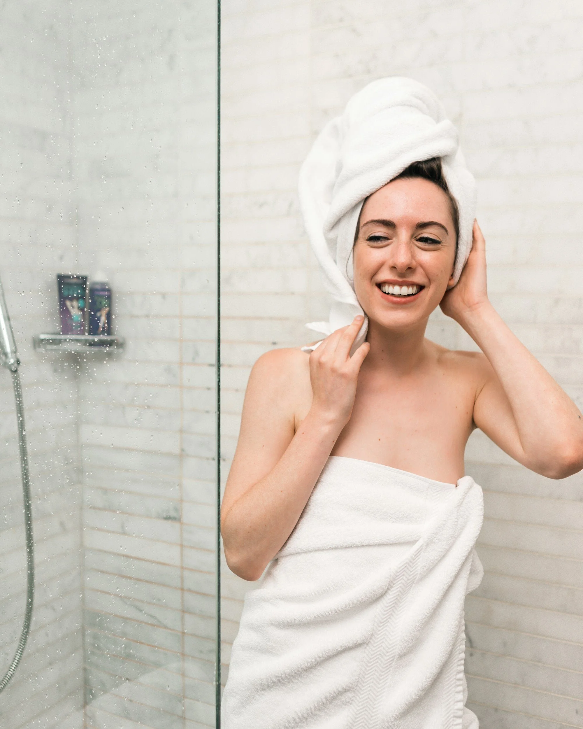 A woman with a towel wrapped around her body and another on her head, standing in a shower, smiling, with a glass shower door and white tiled wall with a small shelf holding shampoo bottles in the background.