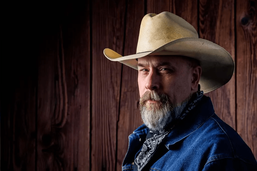 A man with a beard wearing a cowboy hat, denim jacket, and bandana against a wooden background.