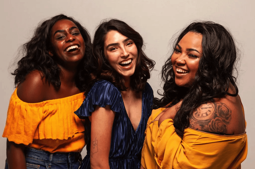Three women smiling and posing together against a plain background, one wearing a yellow off-shoulder top, another in a blue dress, and the third in a yellow dress with tattoos on her arm.