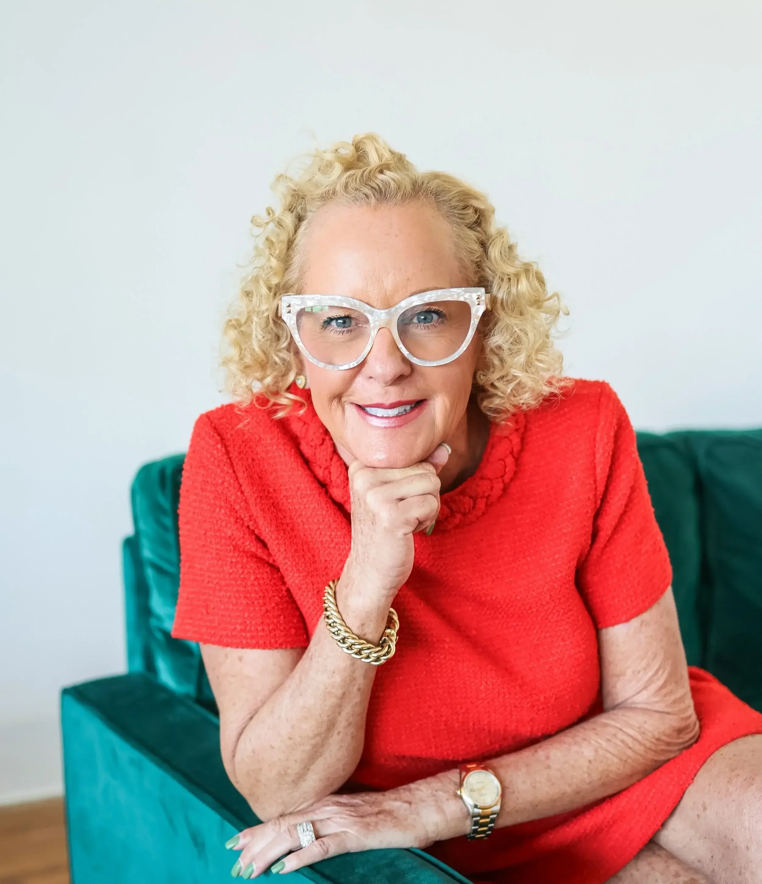 A woman with curly blonde hair and glasses sitting on a green couch, wearing a red dress and gold jewelry, smiling at the camera.