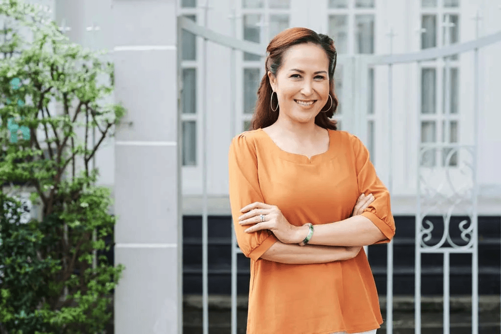 A woman with brown hair and hoop earrings standing outside, smiling, with her arms crossed, in front of a white building with windows and greenery.