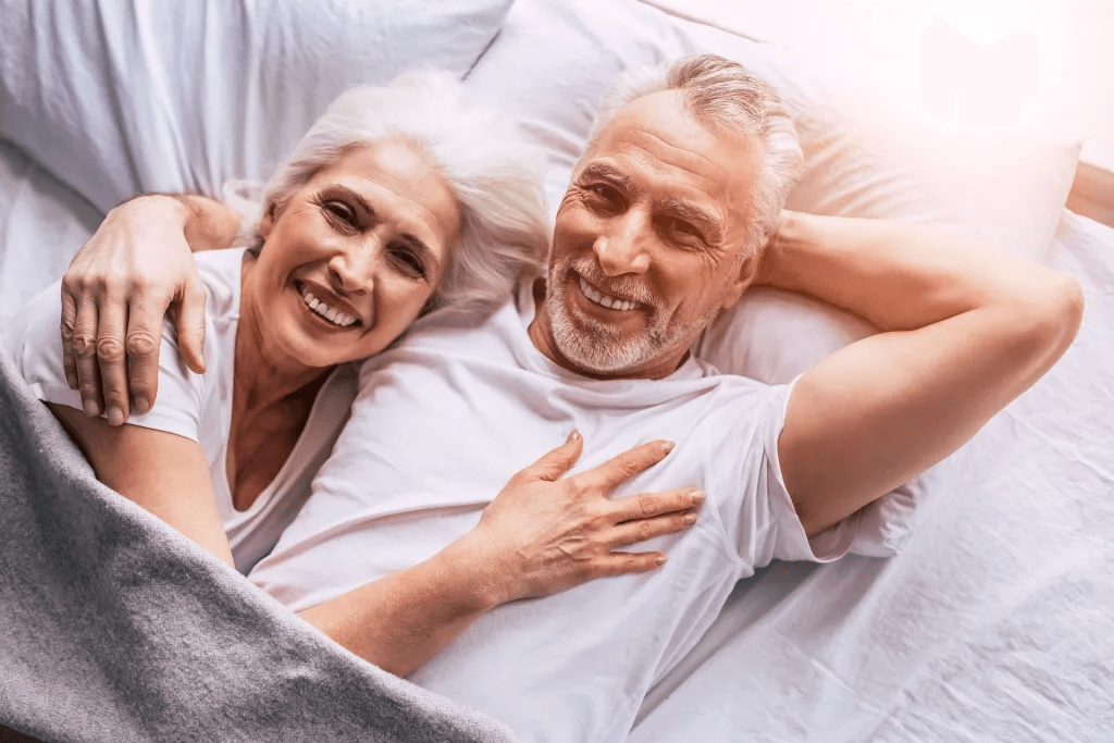 Smiling older couple lying in bed together, cuddling under a gray blanket in bright, natural light.
