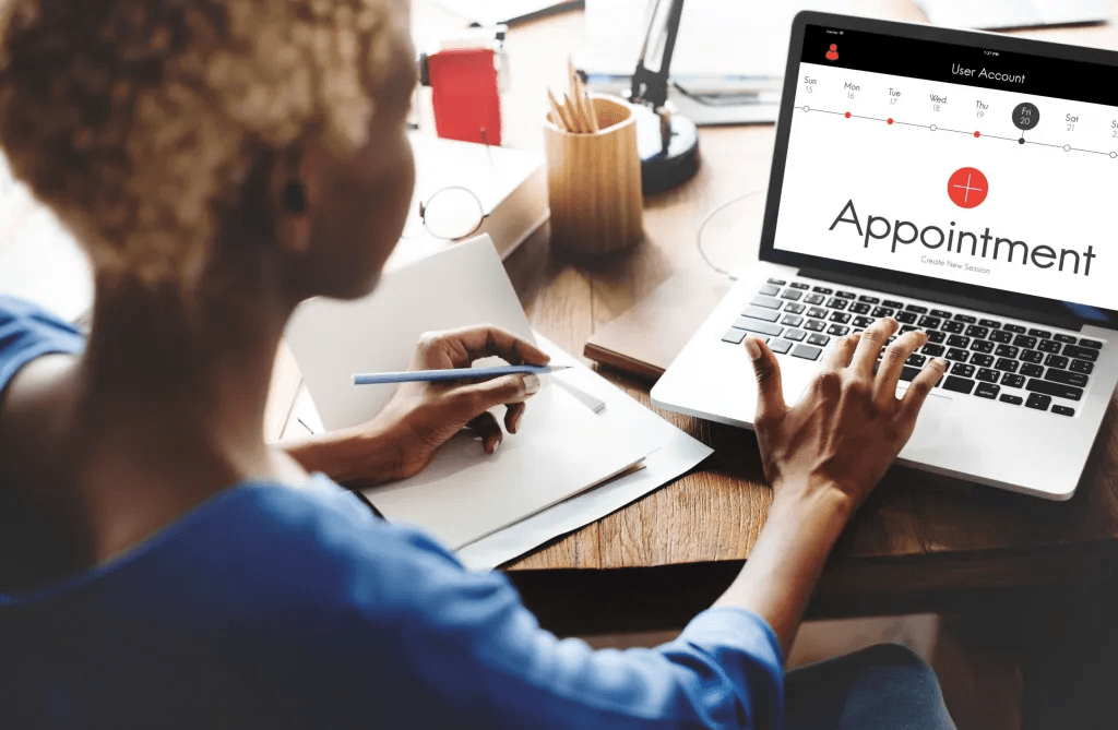 A woman sitting at a desk uses a laptop displaying an appointment scheduling interface, with a notebook and pen in hand, in a well-lit office environment.