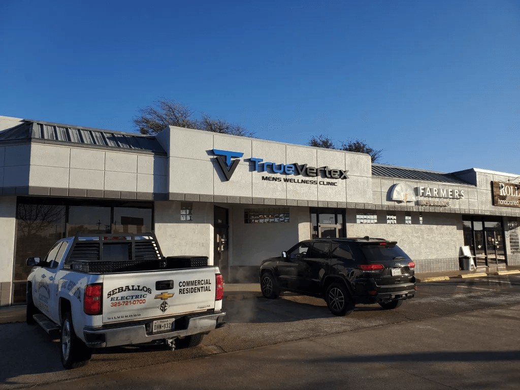 Parking lot in front of a strip mall with signs for TrueVereTx Men's Wellness Clinic, Farmers Insurance, and Roll cosmetics. Two vehicles parked, a white pickup truck and a black SUV. Clear blue sky.