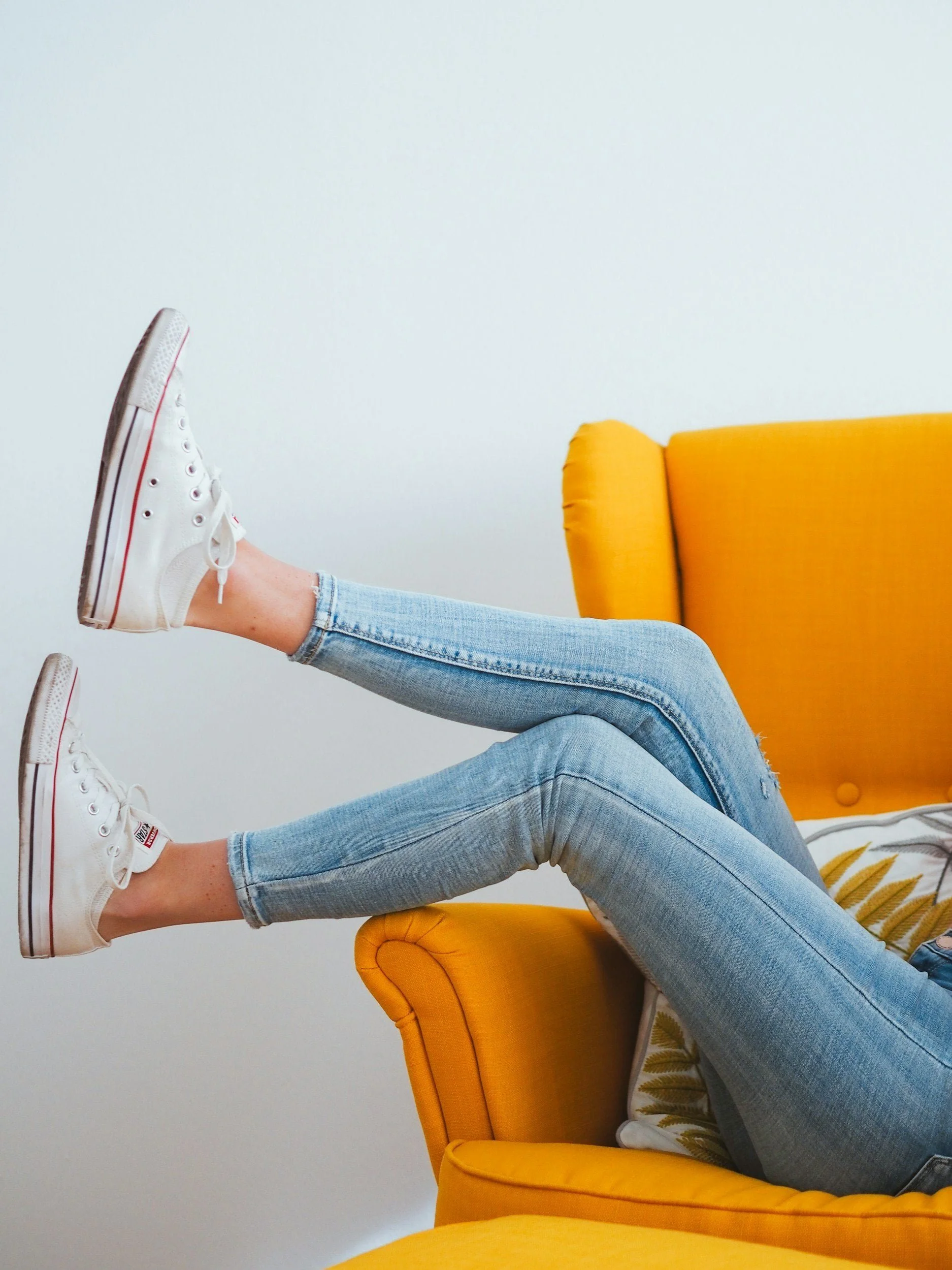 Person lying on a yellow couch with legs raised and wearing white sneakers and light blue jeans.