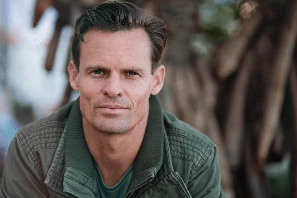A man with short dark hair, blue eyes, and a serious expression, wearing a green jacket, outdoors with blurred trees in the background.