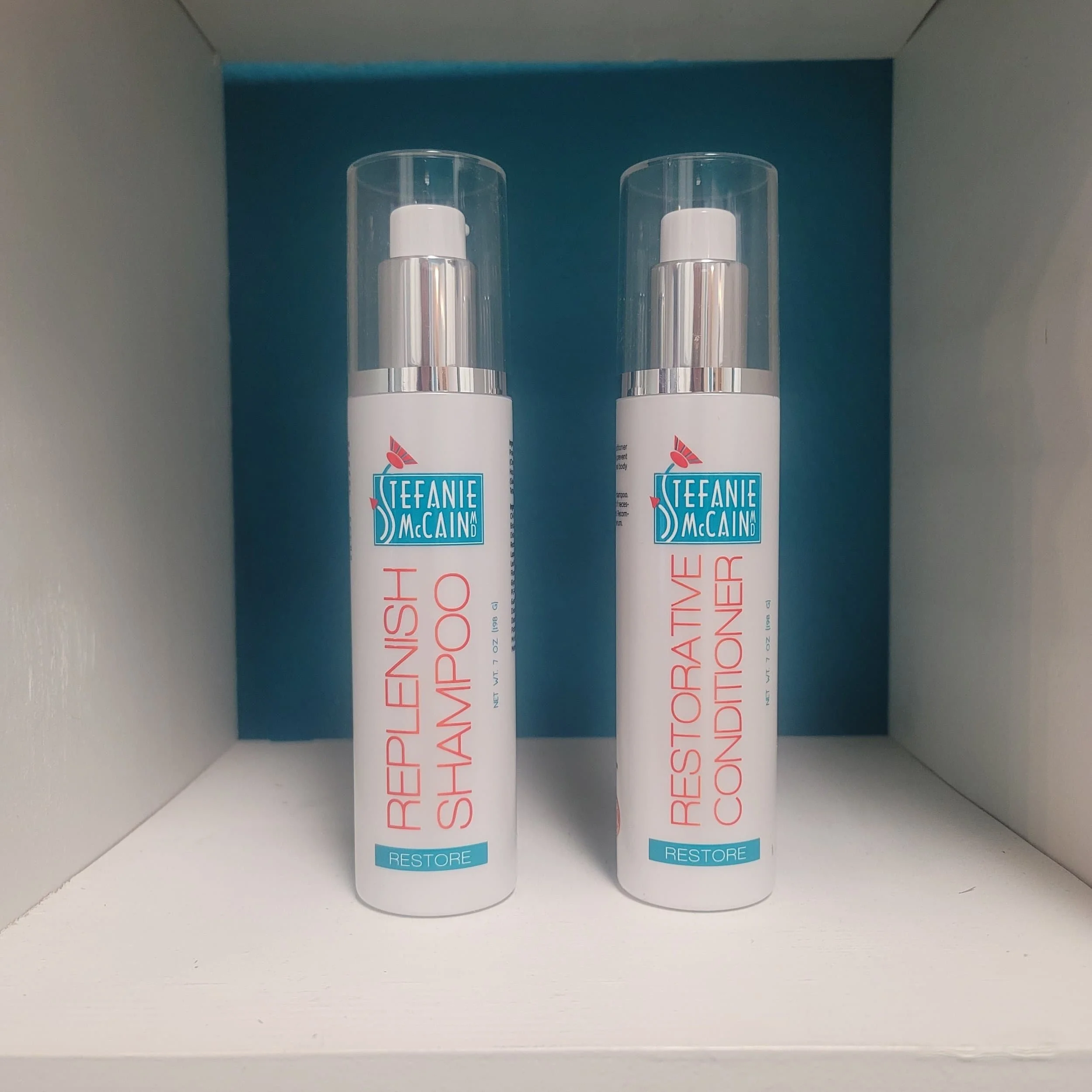 Two bottles of Stefanie McCain Restore hair care products, labeled 'Replenish Shampoo' and 'Rejuvenate Conditioner,' placed on a white shelf with a blue background.
