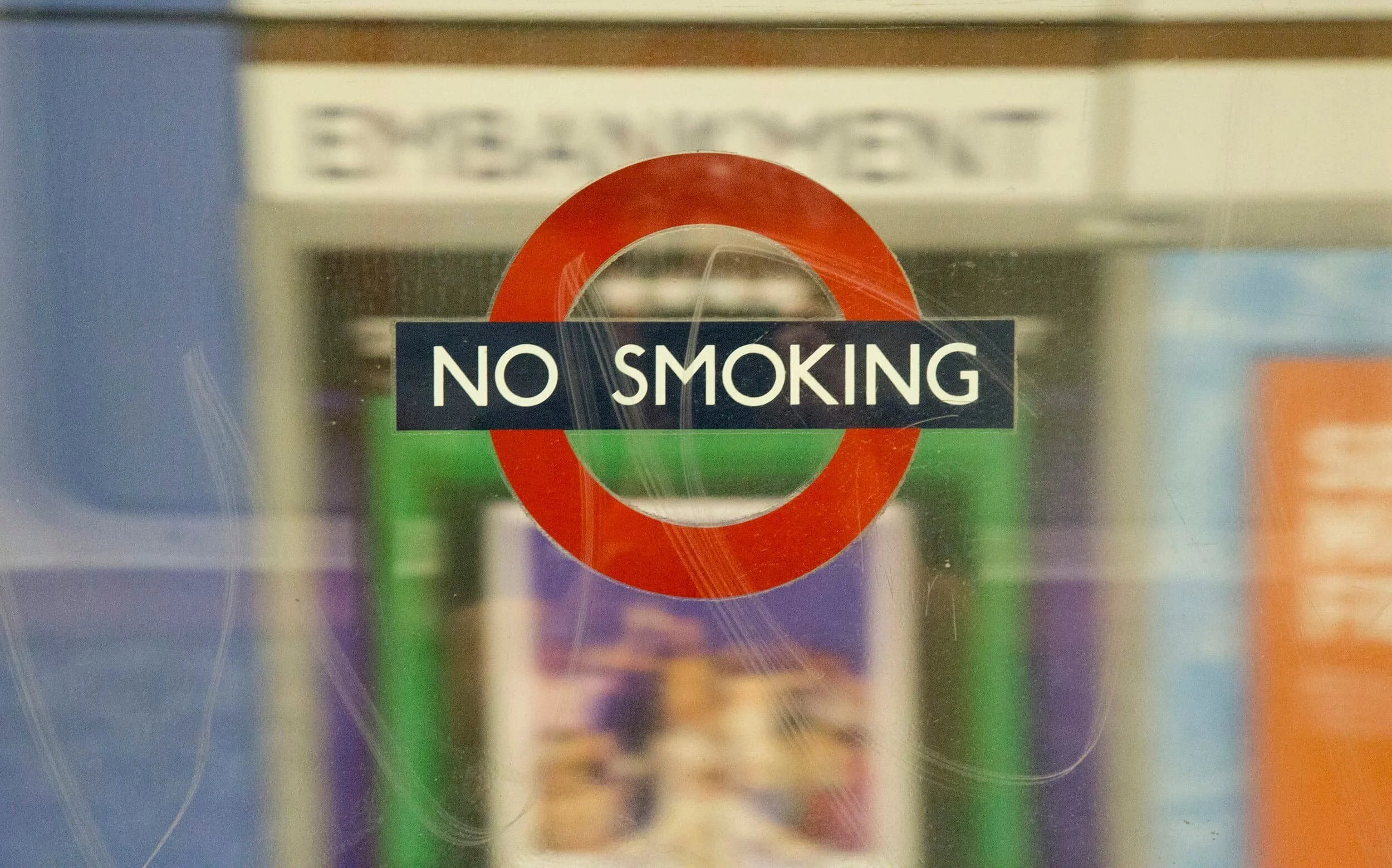 A glass door with a round red and white no smoking sign and a black and white text overlay that reads 'NO SMOKING'. The background shows blurred signs or posters.