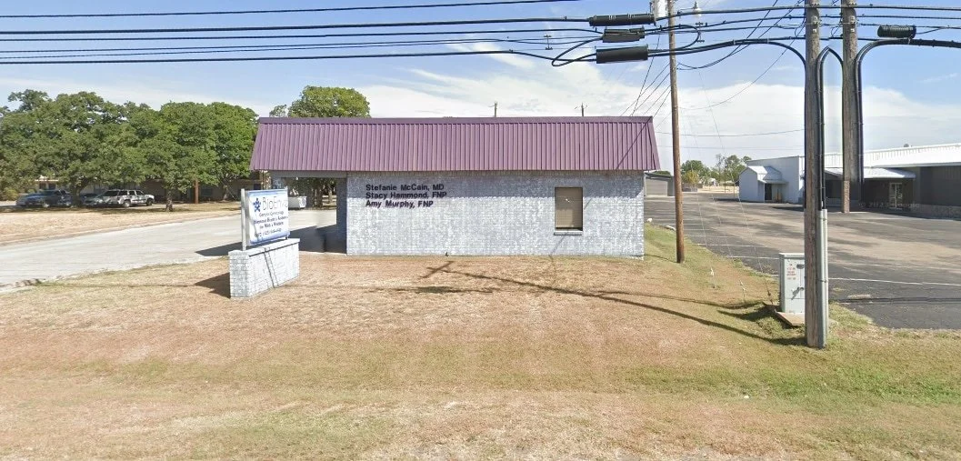 A small building with a purple metal roof and light gray brick exterior, signs indicating medical professionals, and a parking lot to the right.