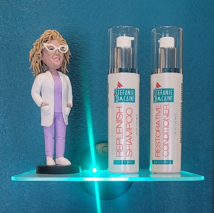 A shelf with a Bobblehead doll of a woman in a white coat and purple scrubs, and two bottles of Teanie McCain shampoo and conditioner with pump dispensers, against a blue wall.