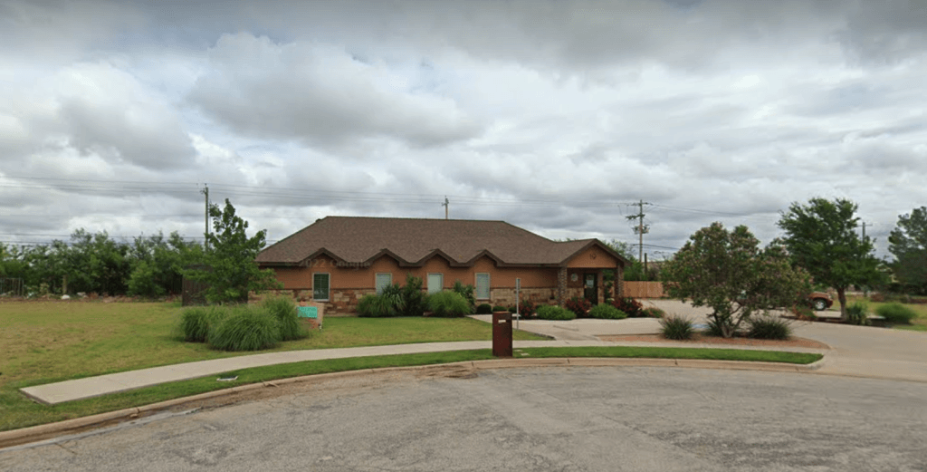 A single-story house with a brown roof and tan exterior walls, surrounded by a well-maintained yard with shrubs and trees, under a cloudy sky, with a parking area in front.