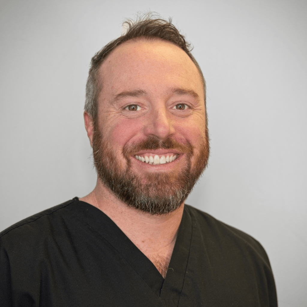 A man with a beard and short hair, smiling, wearing a black medical scrub top, standing against a plain light gray background.