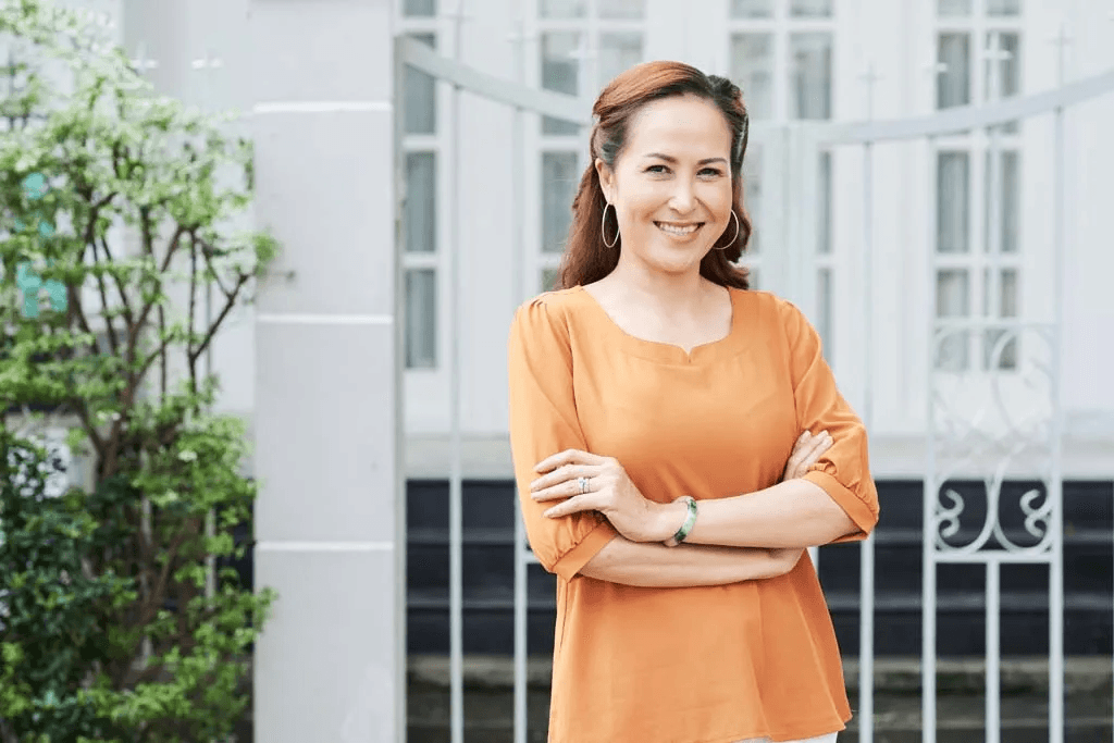 A woman with brown hair wearing an orange top standing outdoors in front of a building with glass windows, smiling with arms crossed.