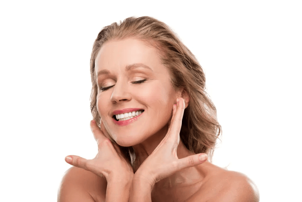 Close-up of a smiling woman with closed eyes, touching her face gently with her hands, against a white background.