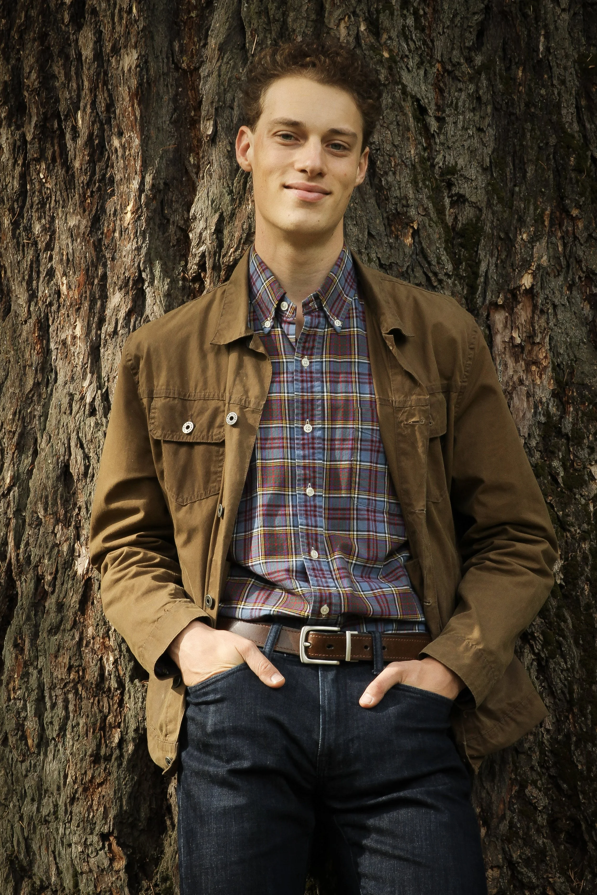 A picture of Sam Kaufman, the co-teacher for grades 1-2 at Portland Forest School.  He is standing in front of a large tree trunk, which takes up the whole background.