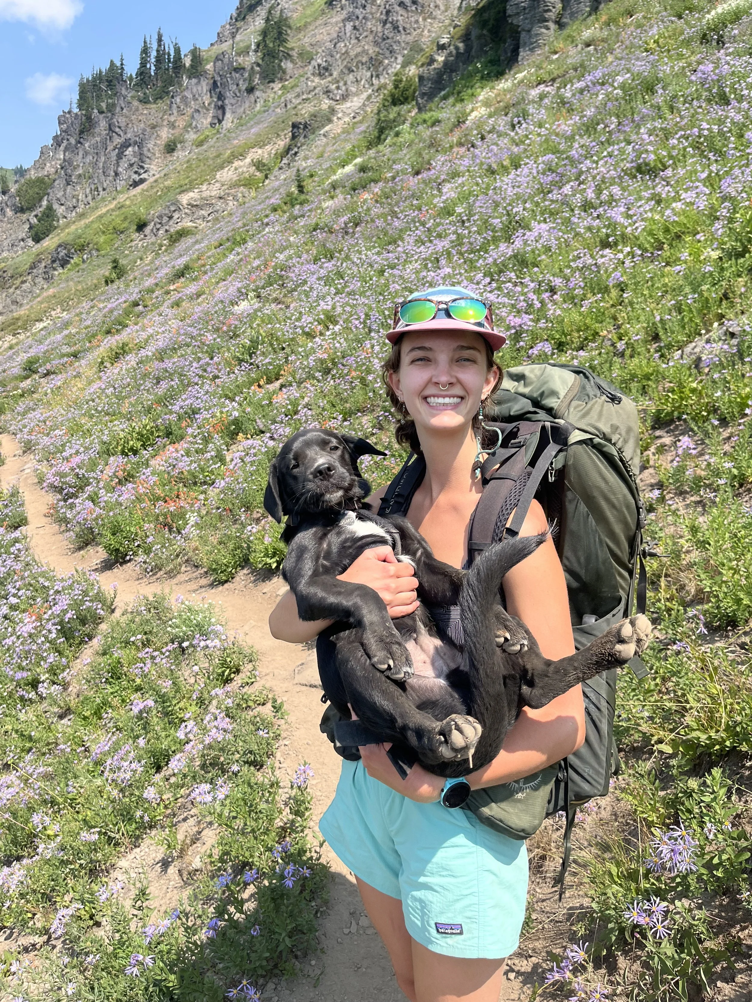 A photo of Grace Van Kirk, the co-teacher of grades 6-8 at Portland Forest School.  Grace is standing on a hiking trail, which runs along a mountain side covered in purple flowers.  She is holder her dog in her arms.