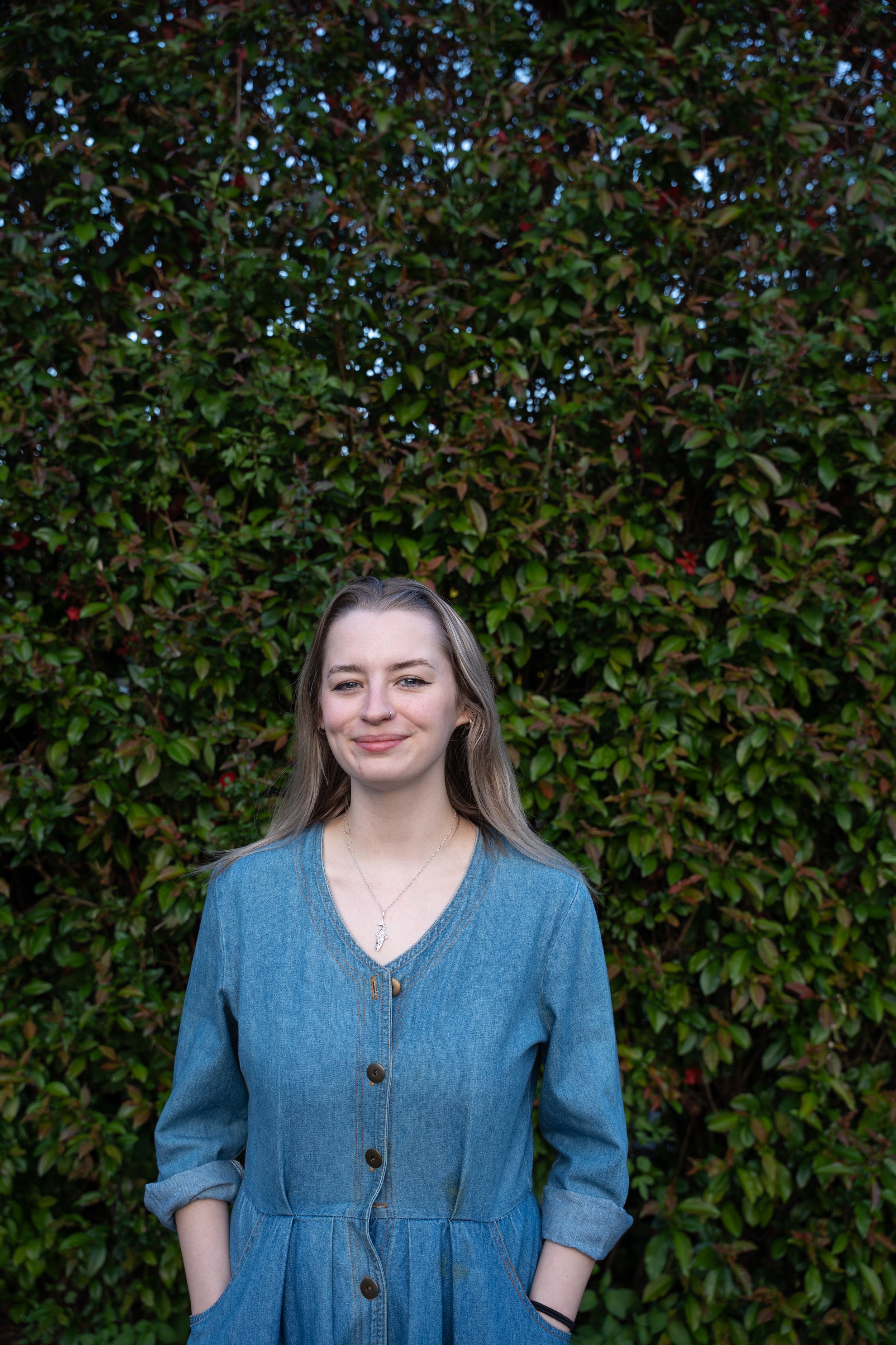 A photo of Hannah Taylor, the art and aftercare teacher at Portland Forest School.  Hannah is standing in front of a large hedge that takes up the full background.