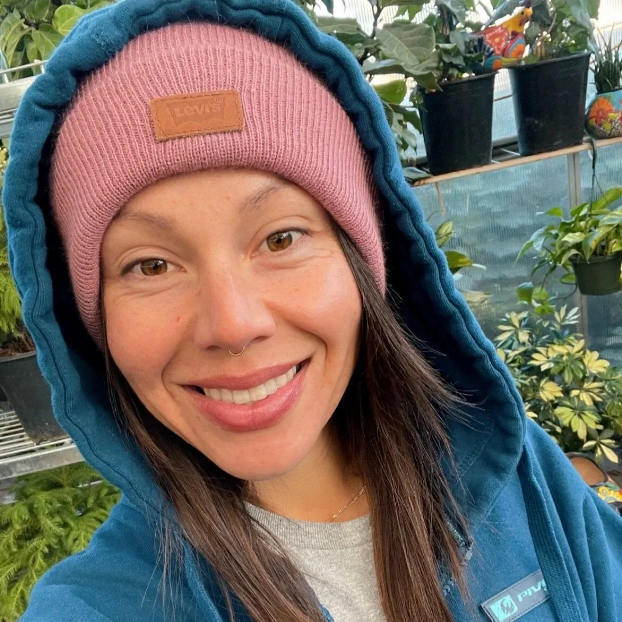A woman smiling outdoors in a greenhouse, wearing a pink beanie, a blue hoodie, and a grey shirt.