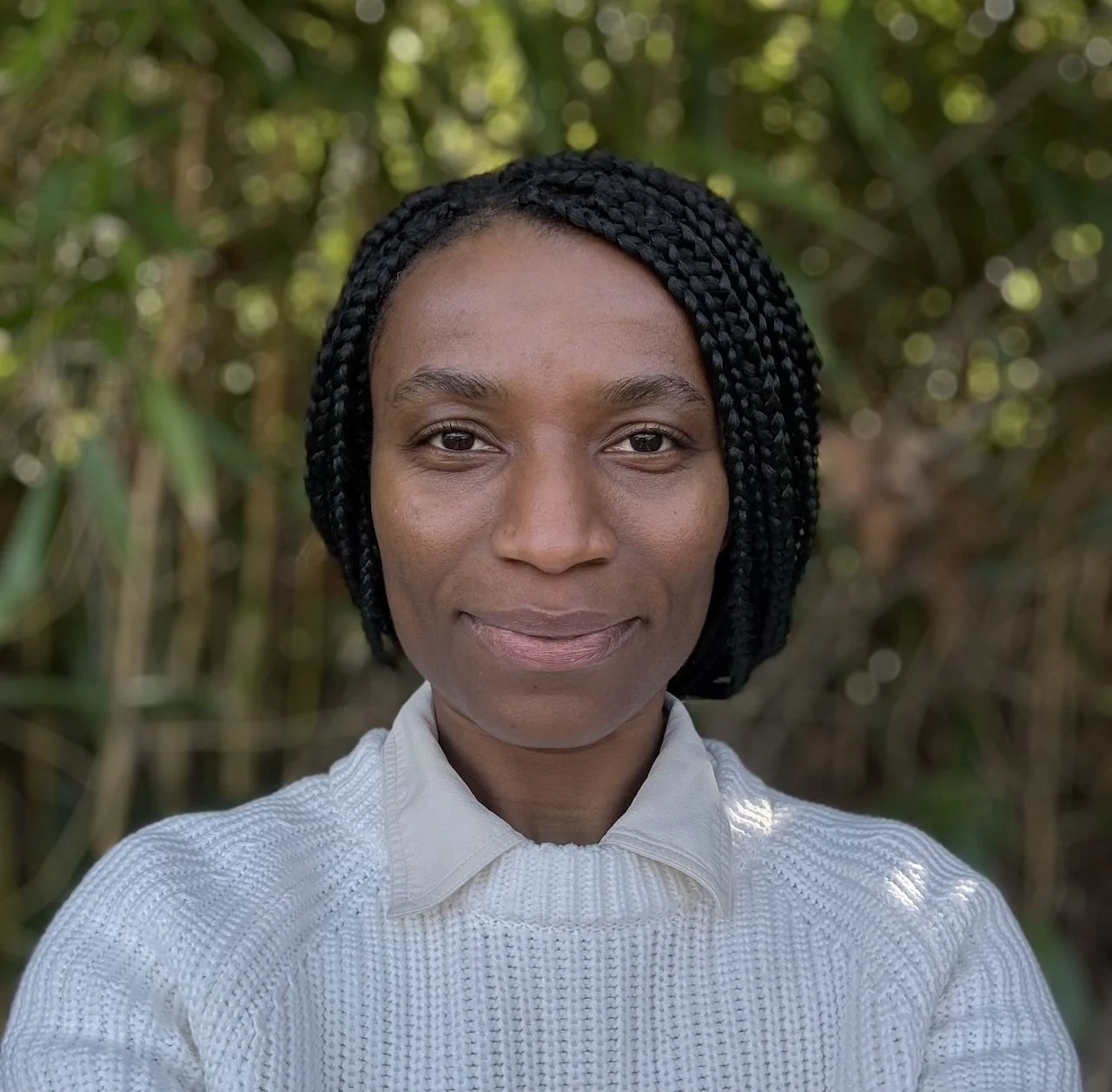 A woman with dark skin and black braided hair stands outdoors in front of greenery, wearing a white textured sweater and a collared shirt, with a gentle smile.