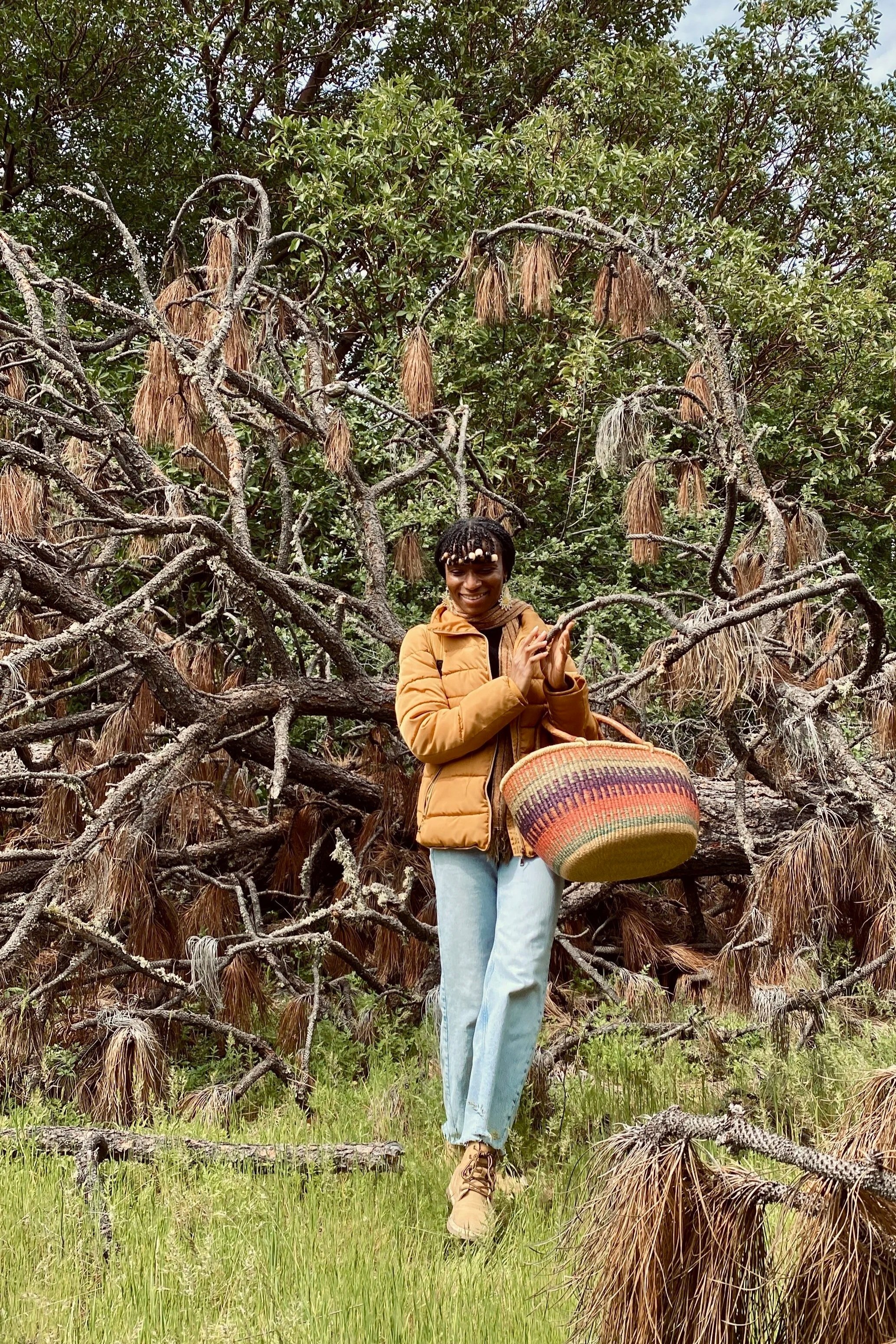 A woman standing outdoors in front of a tree with brown, dried leaves hanging from its branches, holding a woven basket, wearing a tan jacket, light blue jeans, and tan boots, smiling.