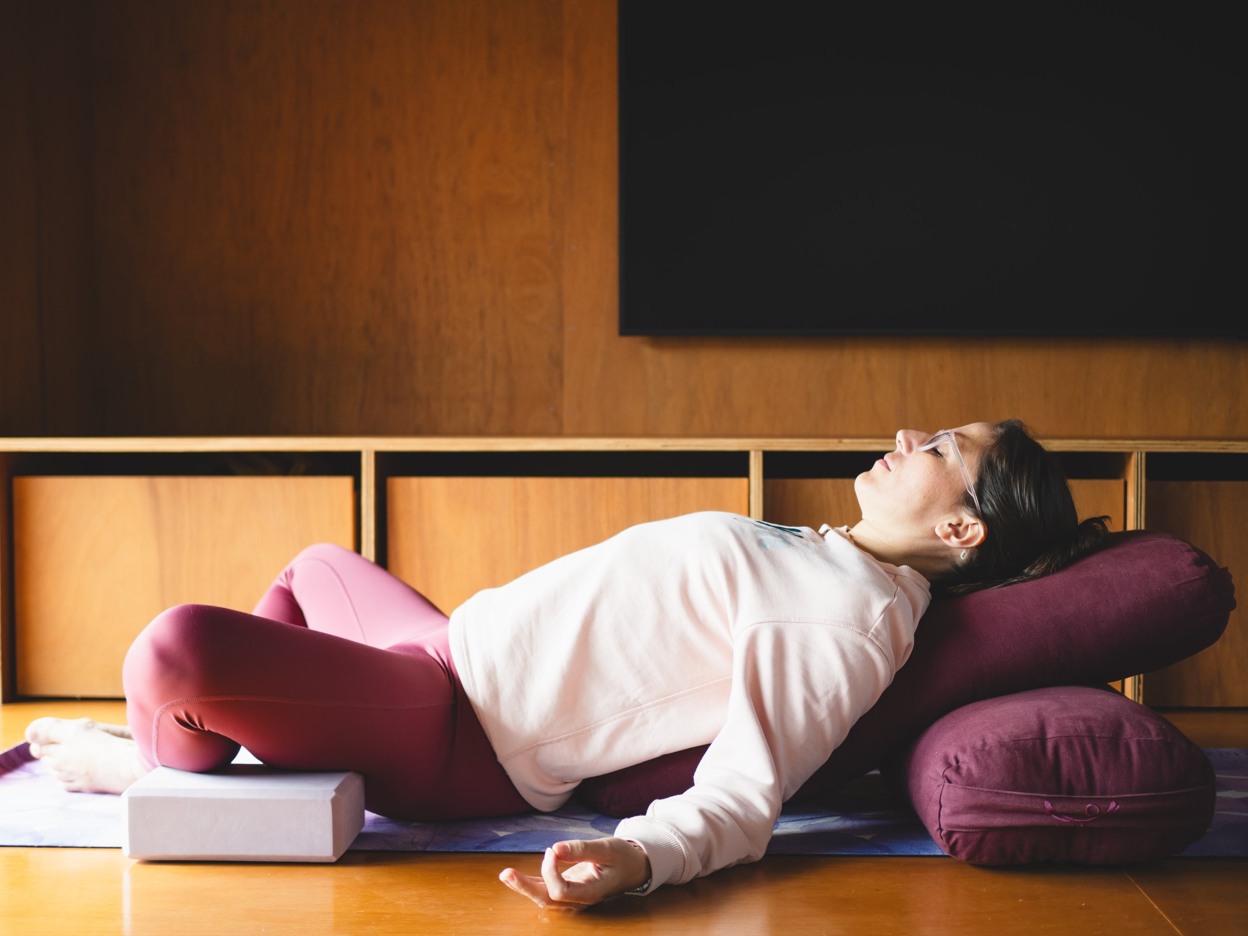 Woman lying onto her back in a supported reclined butterfly position (supta baddha konanasan), wearing a pink jumper and purple tights. Her eyes are closed. She is lying on a purple bolster and has pink blocks supporting her knees.