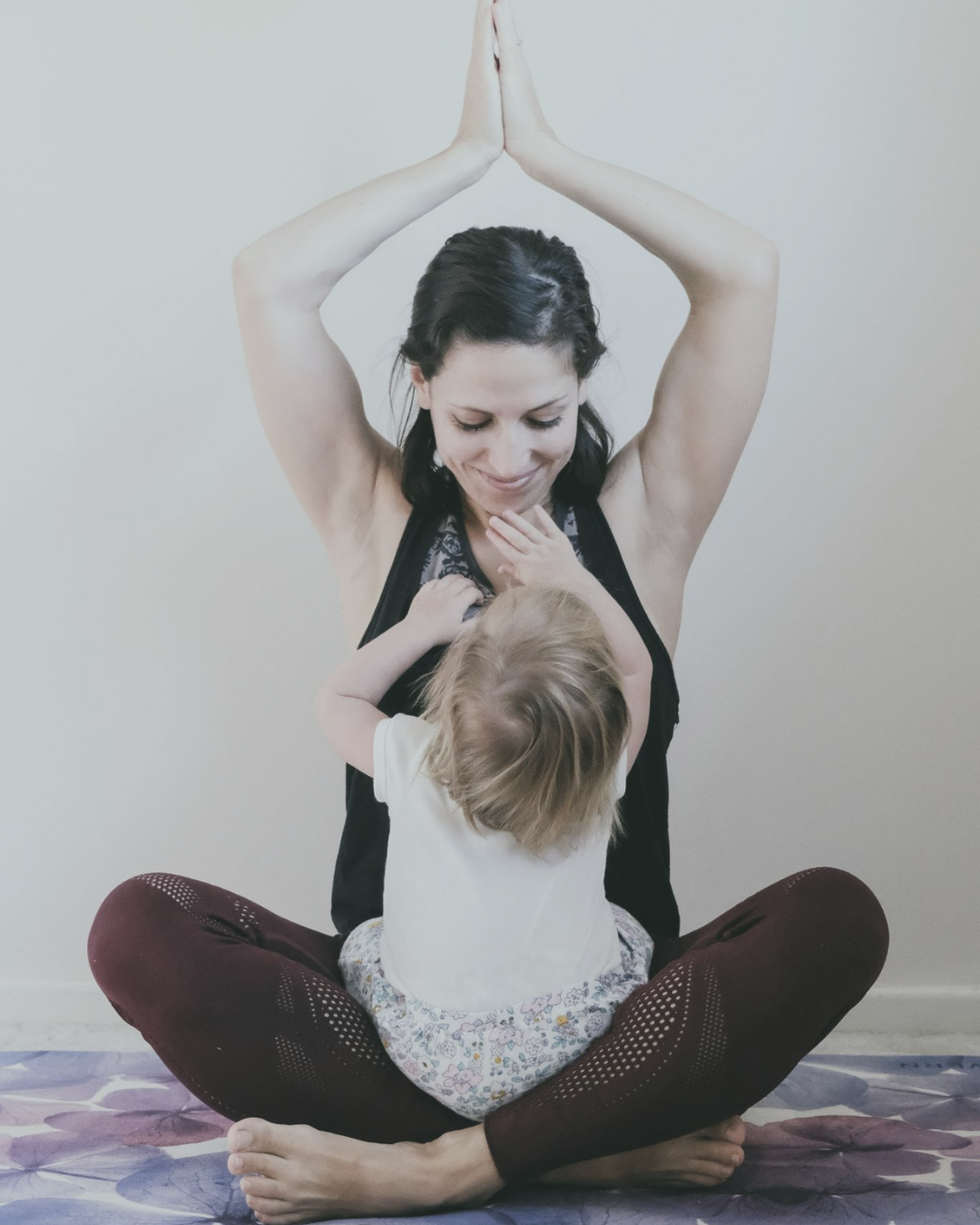 Marion, yoga instructore, showing a seating yoga pose while her baby is sitting on her lap, reaching for her face. She smiled dwon at her baby