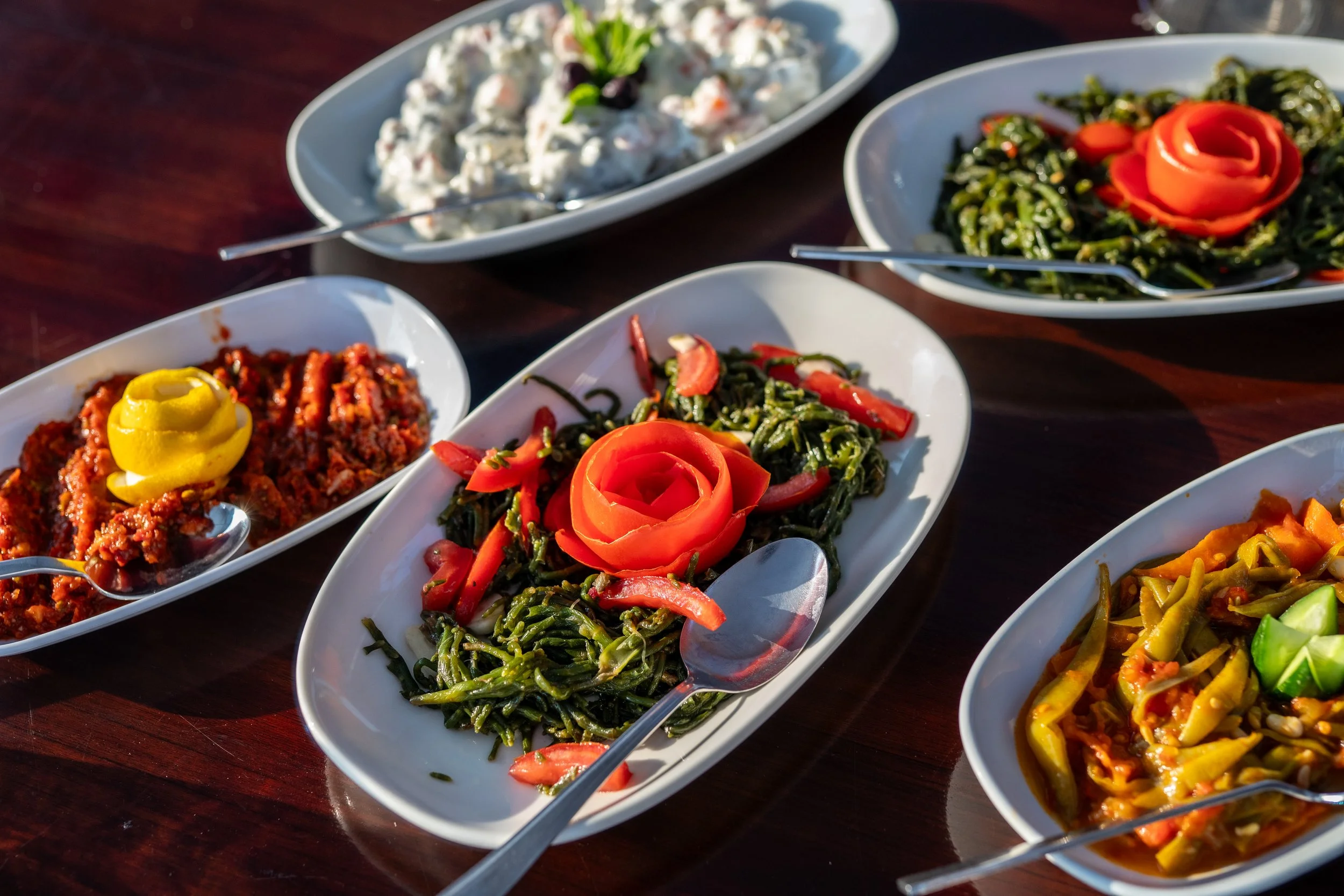 Assorted dishes of salads and pasta on a wooden table, including spinach pasta with tomato rose garnish, and various vegetable salads.