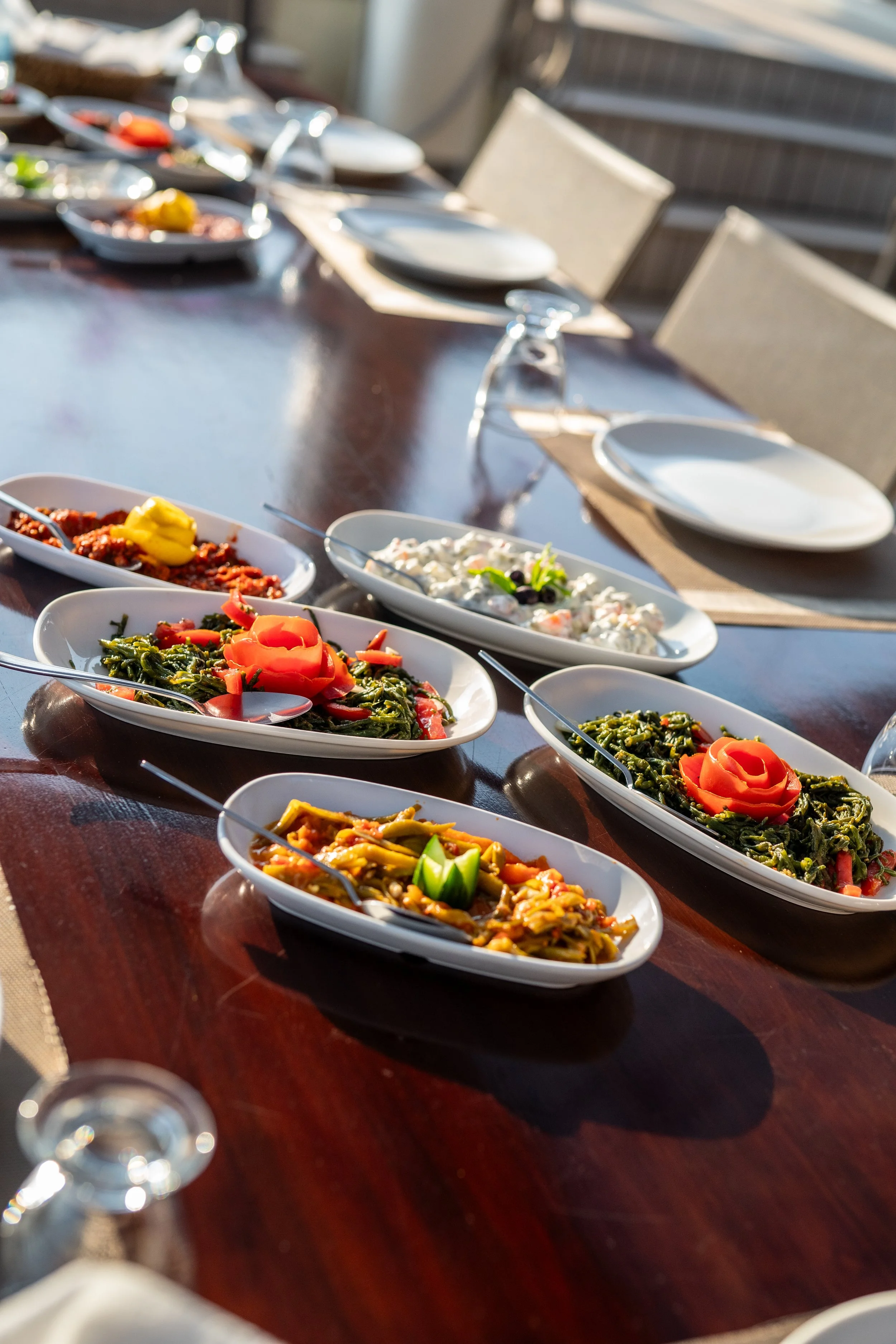 A dining table set outdoors with various dishes of salad and side dishes in white bowls, empty plates, and upside-down glasses.