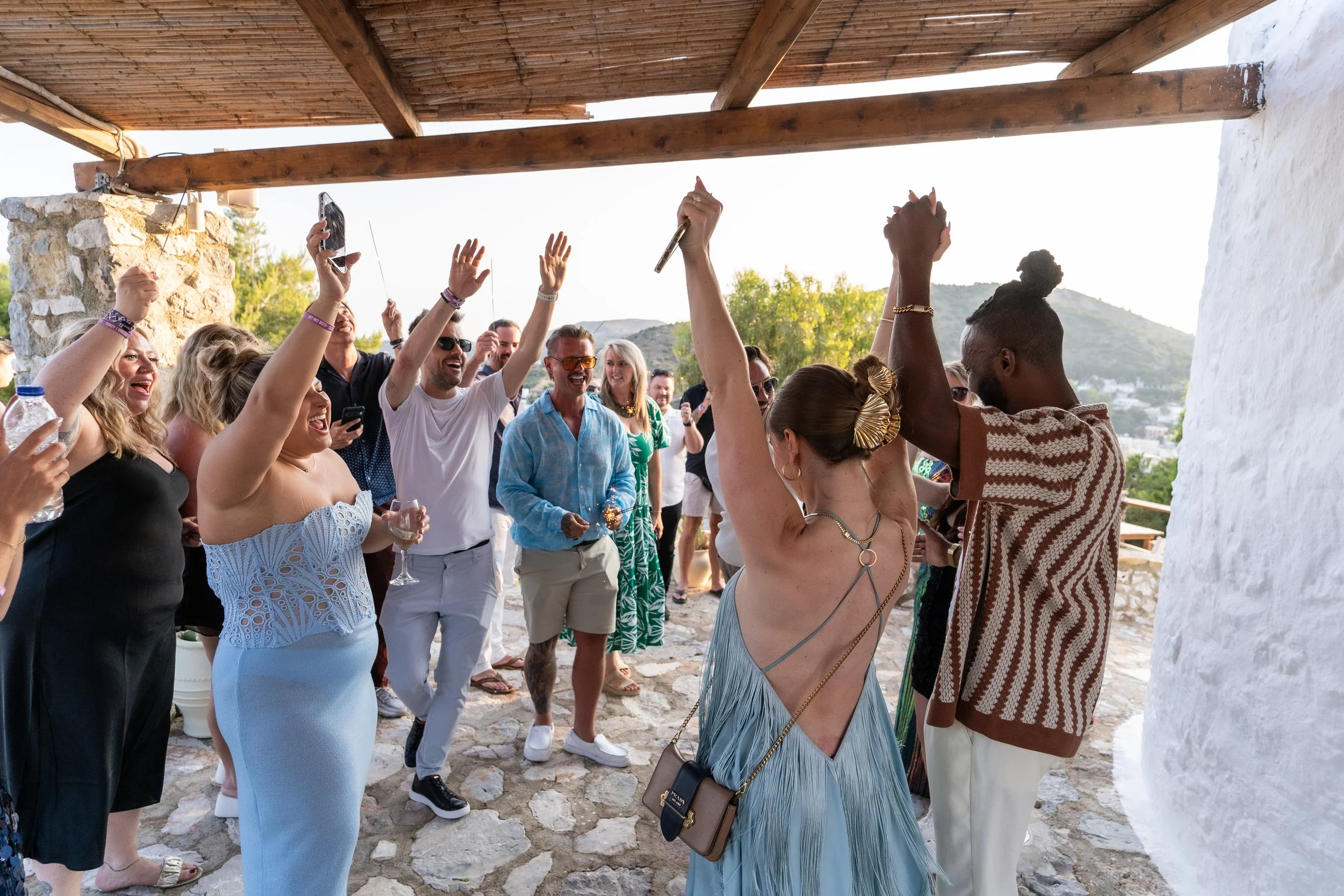 A group of people celebrating at a party outdoors, with some dancing and others raising their hands, under a wooden roof with a scenic background of trees and mountains.