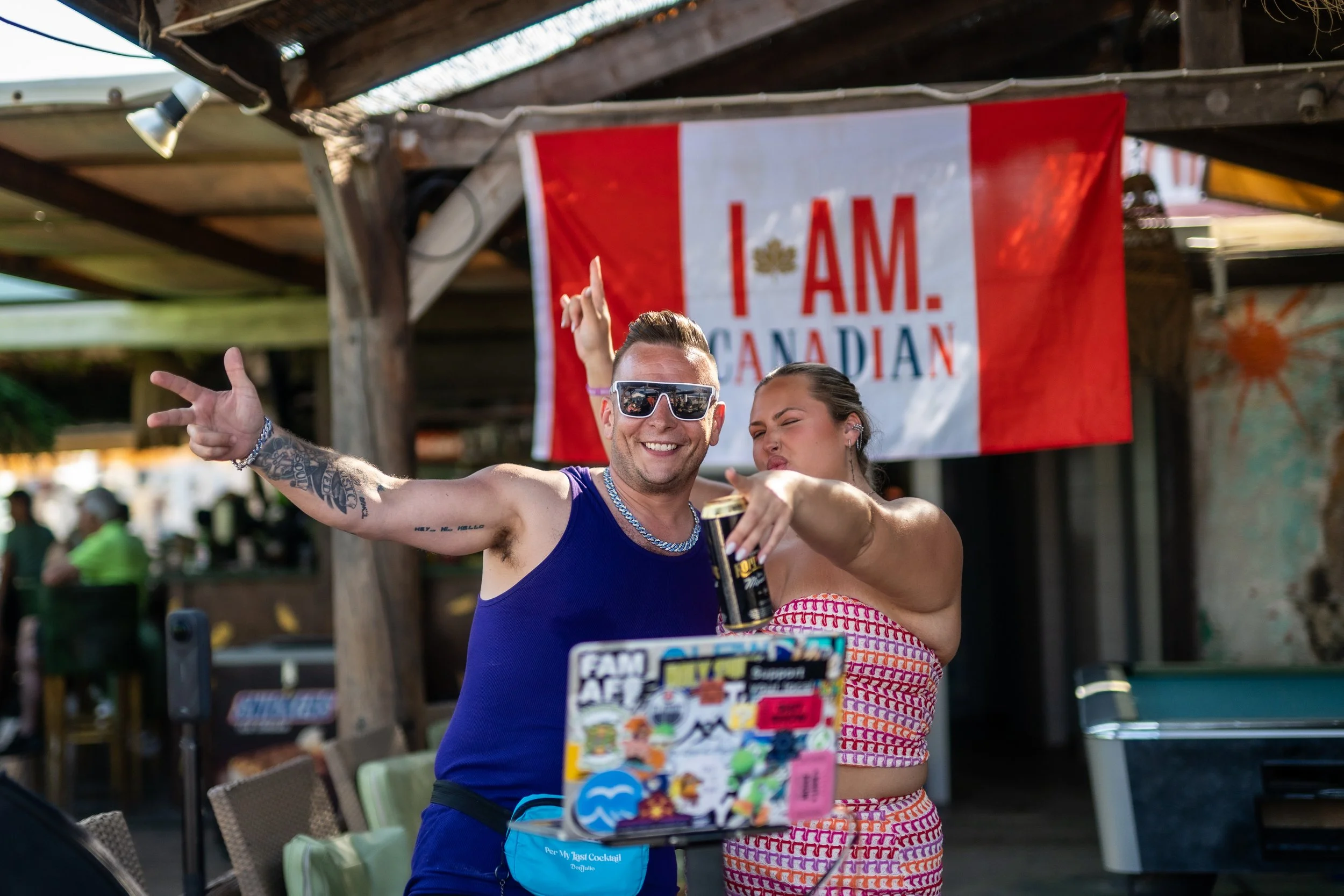 Two people are taking a selfie outdoors under a wooden canopy, with a Canadian flag backdrop that reads 'I AM. CANADIAN'. The man in front is smiling, wearing sunglasses and a blue tank top, and holding a beverage. The woman behind him is making a pl